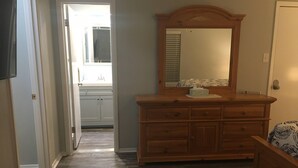 A hallway leading to a bathroom with a wooden dresser, a mirror, and a bed with patterned bedding. - Cozy Modern 1 Bed 1 Bath next to Park! (Van)