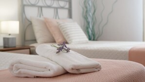 Un lit bien fait, avec une serviette blanche pliée et une fleur violette posée dessus, une table de chevet avec une lampe, et un mur orné d’un motif ondulé.