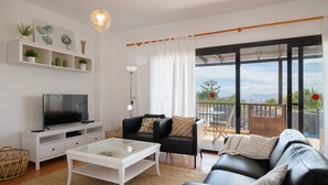 Un salon moderne avec un téléviseur à écran plat, une table basse en verre, un canapé en cuir noir et une console blanche. Il y a un balcon avec vue sur la mer et une terrasse en bois.