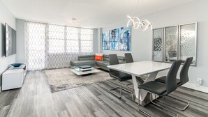 A modern living room with a sofa, a dining table, and chairs. There is a coffee table, a TV, and artwork on the walls. - 2BR 2BA AMAZING NEWLY RENO BEACHFRONT APT W/BEACH VIEW (Hollywood)