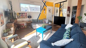 Un salon avec un canapé bleu, une table basse en bois et un fauteuil gris. Une fenêtre offre une vue sur les bâtiments à l’extérieur.