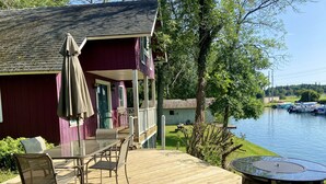 Una terraza de madera con mobiliario exterior, un porche cubierto y vistas a un lago con barcos.