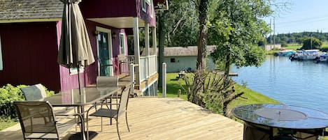 Una terraza de madera con mobiliario exterior, un porche cubierto y vistas a un lago con barcos.