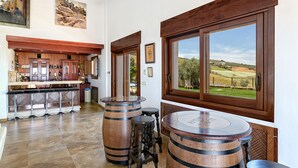 A kitchen with wooden barrels as stools, a bar area with bottles, and a large window with a view of the countryside.