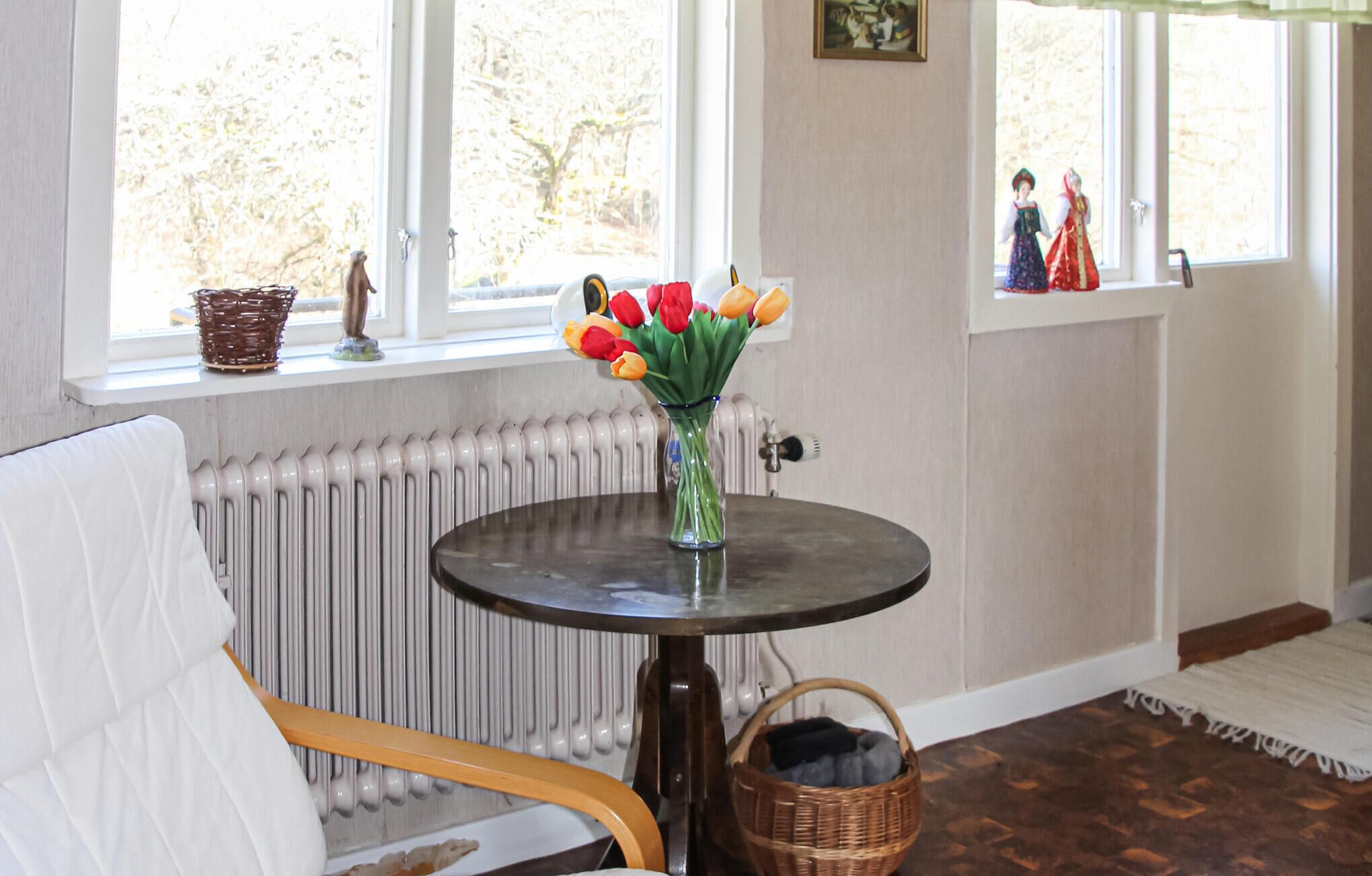 Une pièce avec une table ronde, un panier en osier, un radiateur et une fenêtre donnant sur des arbres.