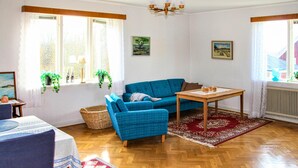 Un salon avec un canapé bleu, une table en bois et un tapis rouge. Il y a deux fenêtres avec des rideaux, un lustre et un tableau au mur.