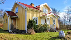 Une maison jaune à deux étages, avec un toit rouge, un porche à l’avant et un petit jardin.