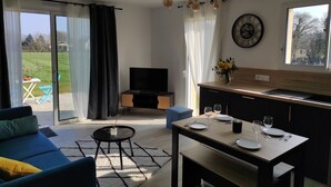 A modern living room with a sofa, a dining table set for two, a TV, and a kitchen area with a clock on the wall.