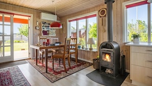 Une pièce chaleureuse avec un poêle à bois, un coin repas avec une table et des chaises, et de grandes fenêtres offrant une vue sur l’extérieur.