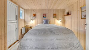 Une pièce chaleureuse aux murs en bois, avec un lit recouvert d’une courtepointe blanche à motifs, une petite table avec une lampe et trois œuvres d’art encadrées accrochées au mur.