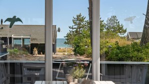 En träveranda med ett bord och stolar, med utsikt över en strand och ett hus med ett tak täckt av tegelpannor.