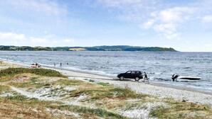 En strand med en bil parkerad vid strandkanten, en båt i vattnet och människor som badar.
