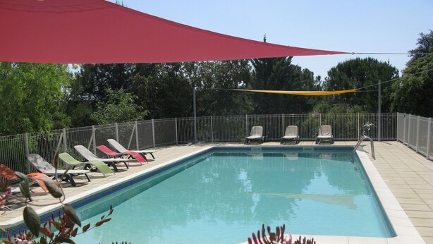 A rectangular swimming pool with a tiled edge, surrounded by a metal fence. There are several lounge chairs along the poolside, and a red shade sail is partially visible above. - House 55m2, large swimming pool (BUZIGNARGUES)