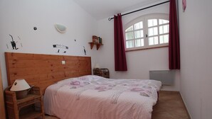 A bedroom with a wooden headboard, a bed with a floral bedspread, a wicker side table with a lamp, a small shelf with books, and a window with curtains. - House 55m2, large swimming pool (BUZIGNARGUES)