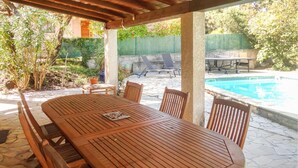 Taula de fusta amb caires sota una pèrgola, una piscina i un pati de pedra.