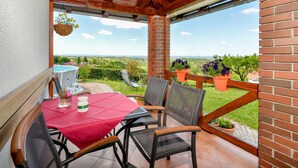 Une terrasse couverte avec une table et des chaises, un pot de fleurs suspendu et une vue sur la campagne.