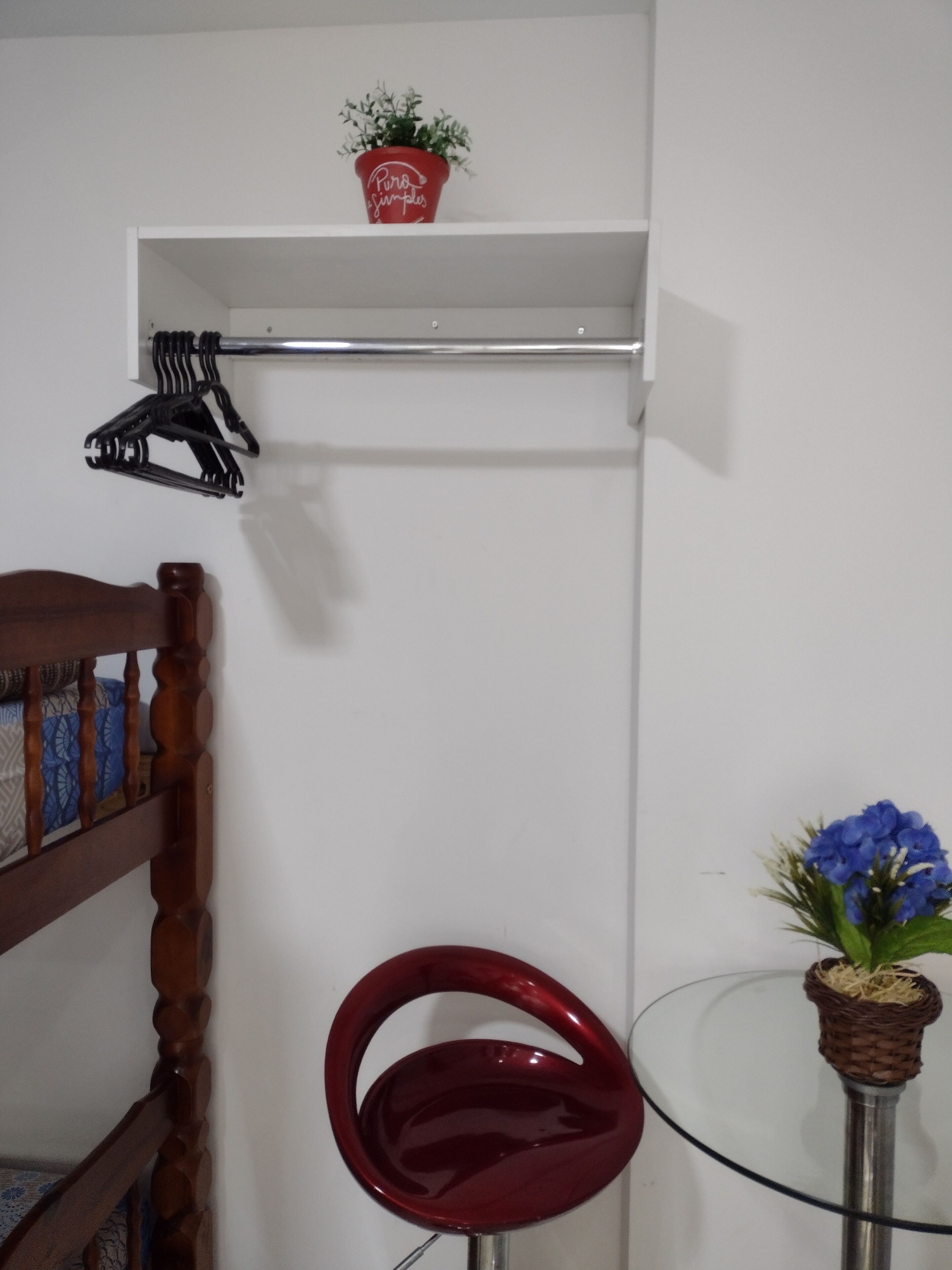 Ein Zimmer mit einem Holzbstuhl, einem roten Sessel, einem kleinen Tisch mit einer blauen Blumenvase und einem Regal mit einem roten Topf und einem schwarzen Kleiderbügel.