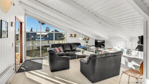 Een ruime woonkamer met een grote leren bank, een kleinere bank, een salontafel en een eettafel. Er staat een televisie op een kast, een lamp en een raam met uitzicht op het exterieur.