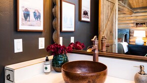 A bathroom with a rustic design, featuring a large copper sink, a green vase with red flowers, and framed pictures on the wall. - Beautiful Log Cabin in Paradise Valley, Montana (Livingston)
