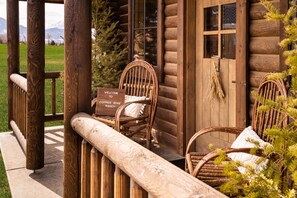 A rustic log cabin with two wicker chairs on the porch, a welcome sign, and a view of the mountains.