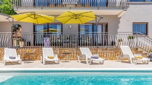 Zona de piscina amb gandules blanques, ombrelles grogues i una paret de pedra.