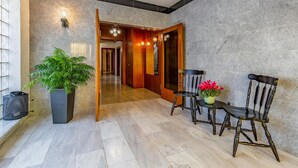 Un passadís amb una porta de fusta que dona a una habitació, una planta en test i dues cadires amb una taula.