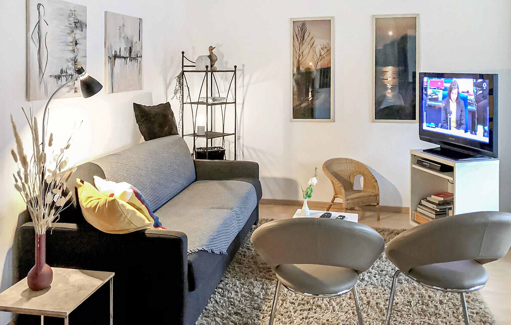 Un salon moderne comprenant un canapé, deux fauteuils, une table basse et un téléviseur. Les murs sont ornés de tableaux abstraits, et une étagère décorative abrite des plantes.
