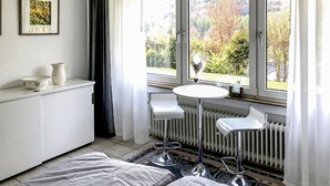 Une petite pièce lumineuse, avec un radiateur blanc, une table blanche et un banc blanc. Il y a une fenêtre donnant sur des arbres et un jardin.