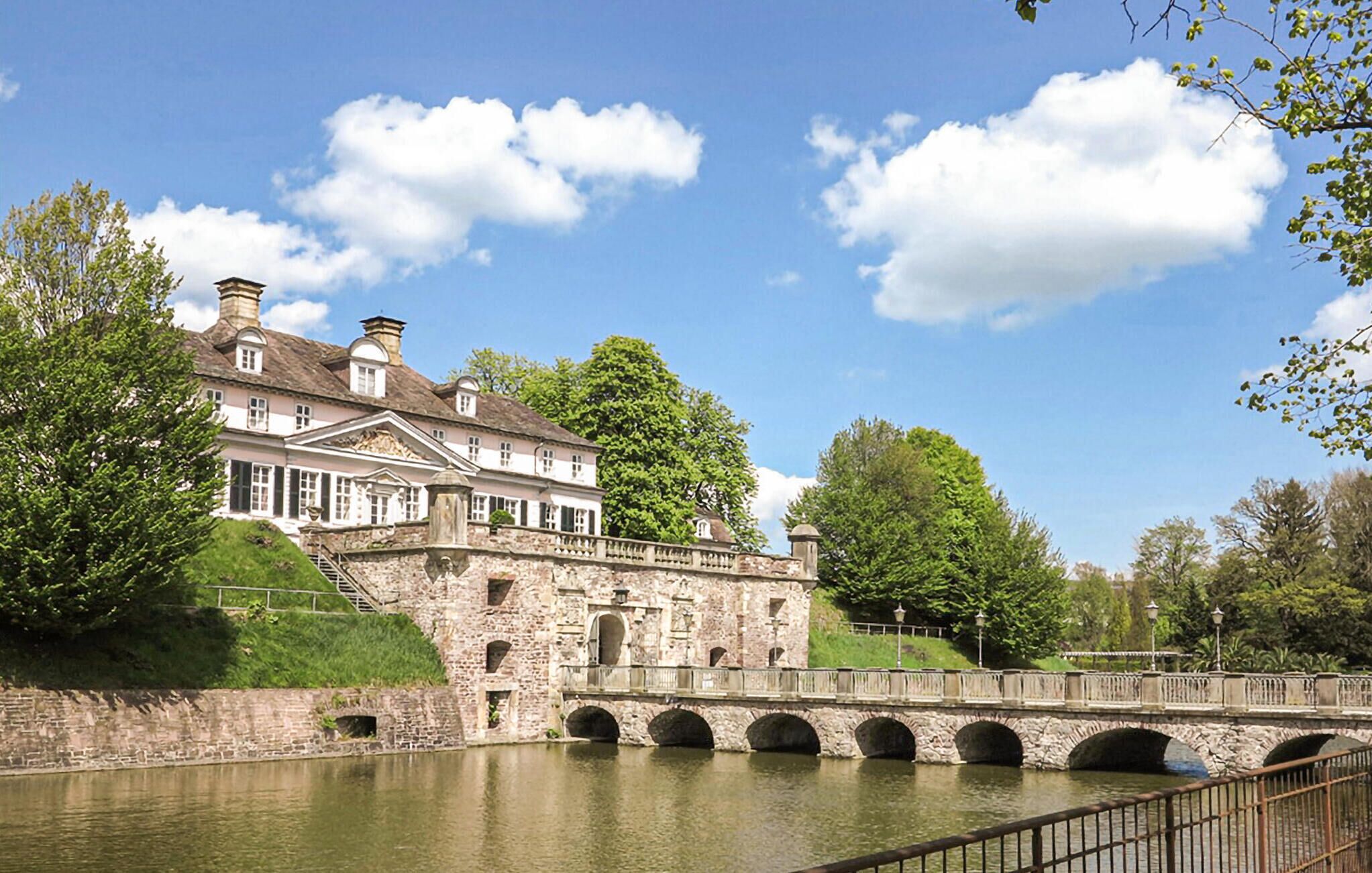 A historic stone building with a bridge over a calm river, surrounded by lush greenery.