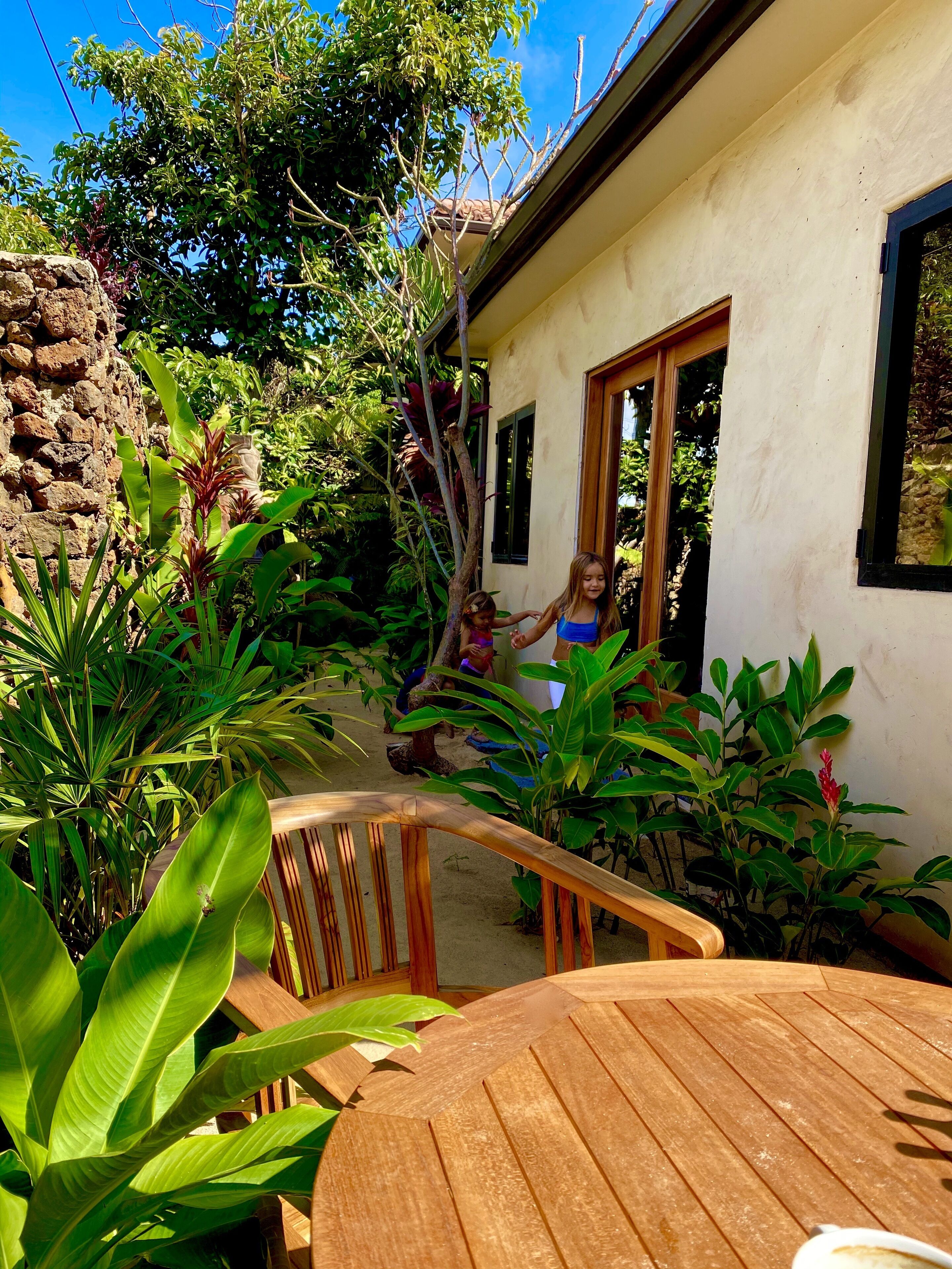 Une table et des chaises en bois se trouvent dans un espace jardin, avec un bâtiment blanc et un mur de pierre en arrière-plan.