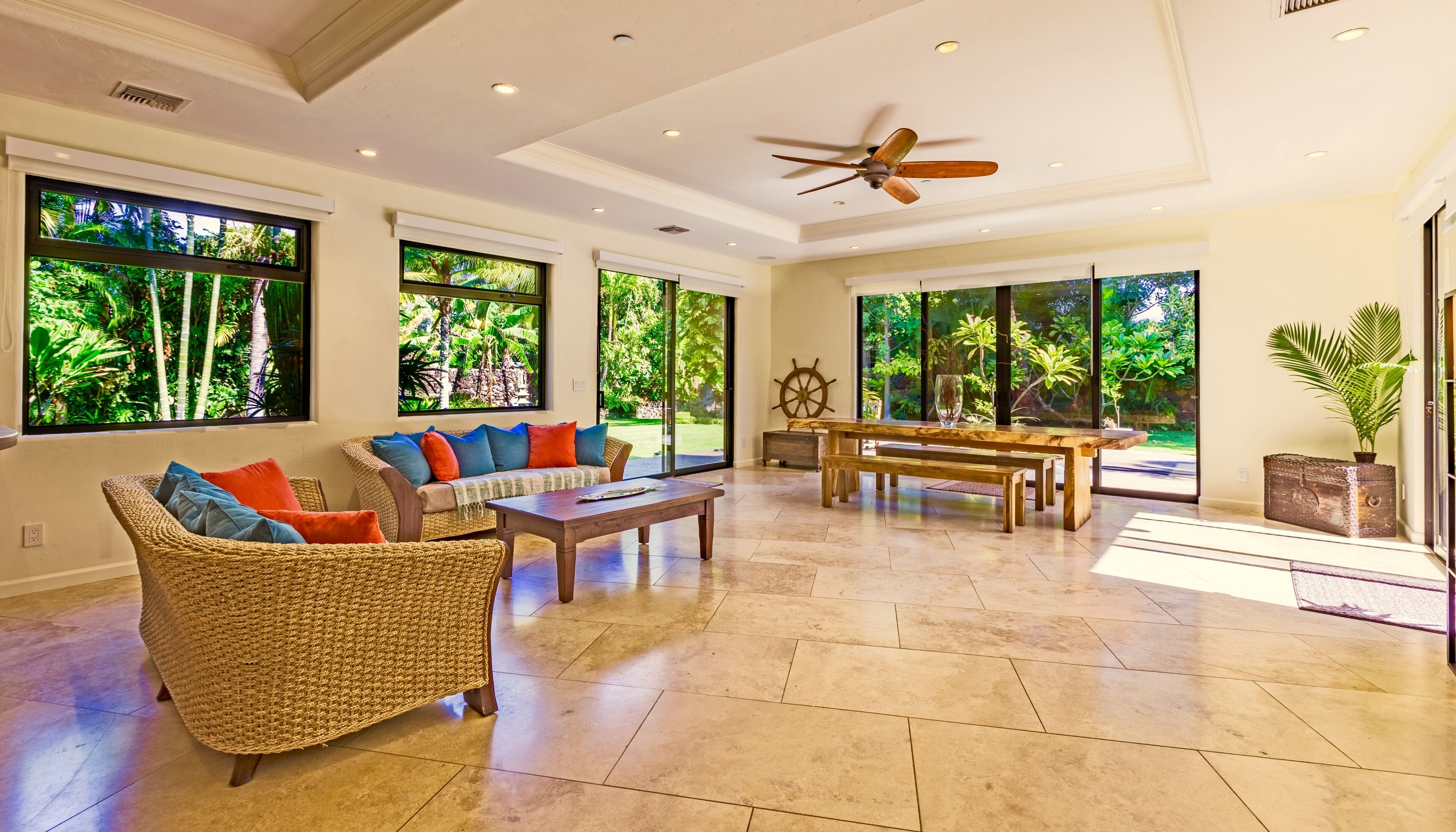 A spacious living area with a large window, wicker furniture, a wooden table, and a ceiling fan.