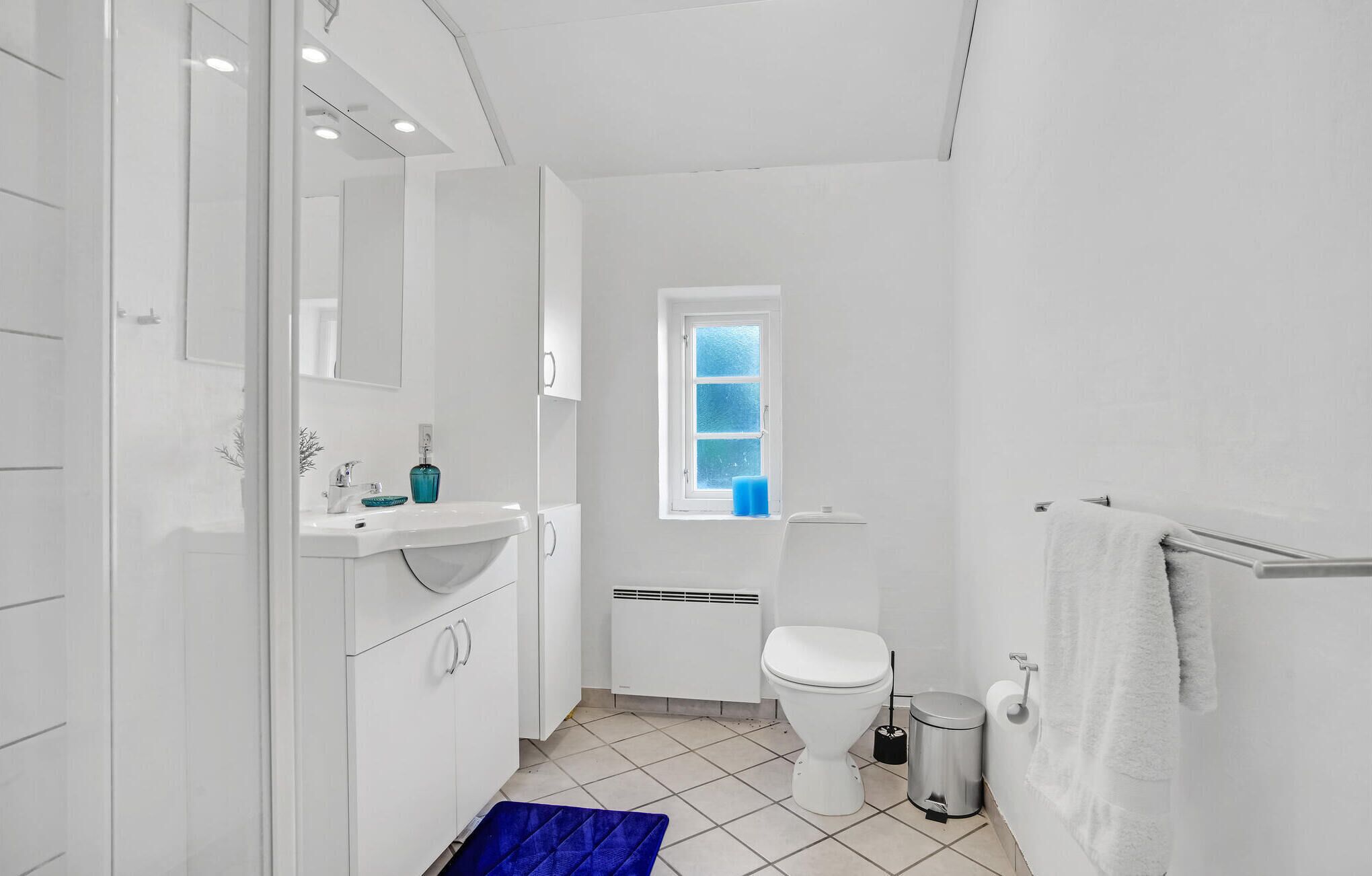 A modern bathroom with a white sink, mirror, and toilet. There is a window with frosted glass, a blue mat, and a white towel rack.
