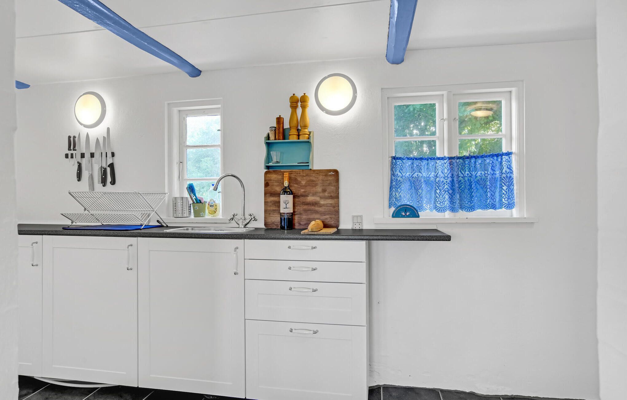 A kitchen with white cabinets, a black countertop, and a window with blue curtains. There are knives hanging on the wall, a sink, and a wine bottle on the counter.