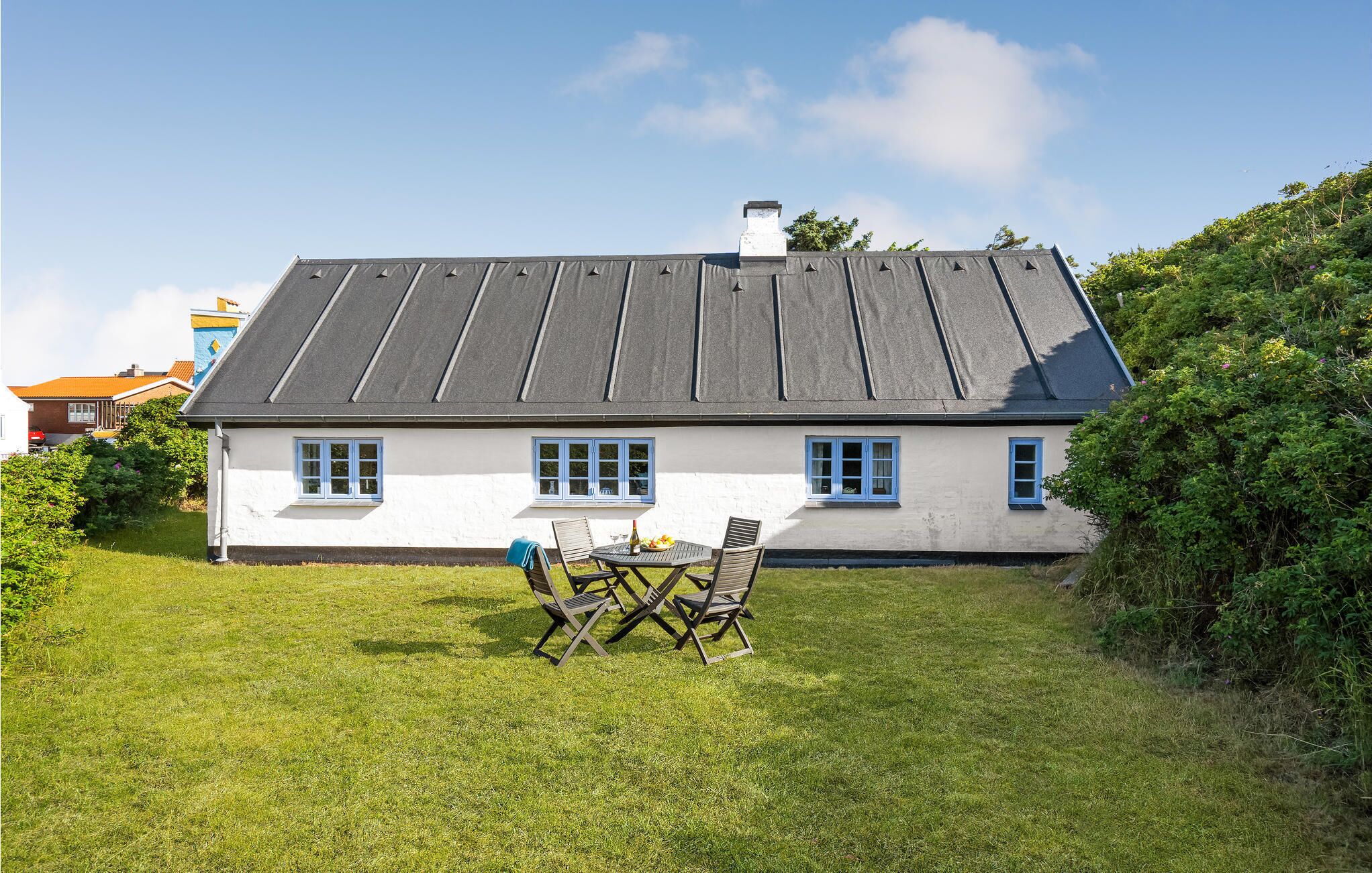 Ett hus med ett grått tak, vita väggar och blå fönsterramar. Framför huset finns en gräsmatta med ett bord och några stolar.