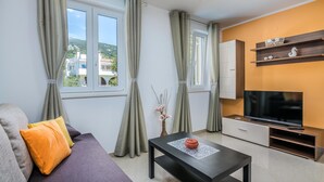 A modern living room with a sofa, a coffee table, a TV, and a view of buildings through the window. - Stunning apartment in Baska with WiFi (Baska)