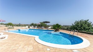 A rectangular swimming pool with a curved edge, surrounded by a tiled deck. There are lounge chairs and umbrellas, and a view of the landscape in the background. - Stunning home in Ragusa (Ragusa)
