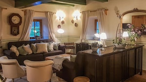 Una sala de estar acogedora con un sofá marrón oscuro, dos sillones blancos, un taburete de madera y una mesita con una lámpara. Un espejo grande y apliques de pared proporcionan iluminación.