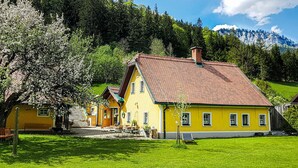Una casa amarilla con techo rojo, rodeada de césped verde y árboles, con montañas al fondo.