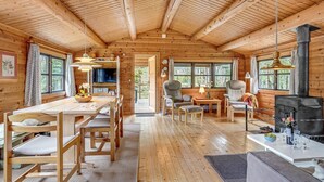 A cozy cabin interior with a dining area, two armchairs, a sofa, a fireplace, and large windows offering a view of the outdoors.
