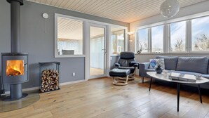A modern living room with a wood-burning stove, a comfortable seating area, and large windows offering a view of the outdoors. - 3 bedroom stunning home in Nexø (Nexø)