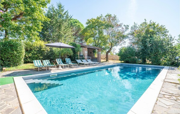 Une piscine rectangulaire à l’eau d’un bleu limpide, entourée d’une terrasse en pierre. On y trouve des chaises longues avec des coussins à rayures et un petit espace salon couvert, avec une table et des chaises.