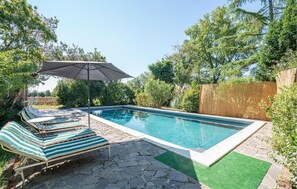 Une piscine rectangulaire bordée de pierres, entourée d’une terrasse également en pierre. On y trouve des chaises longues et un grand parasol qui offre de l’ombre.