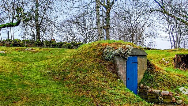 Edifici de pedra rústic amb una porta blava, envoltat de vegetació exuberanta i un mur de pedra en un paratge natural.