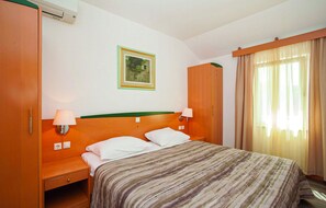 Une chambre d’hôtel avec un lit, deux tables de chevet, une petite table, une lampe fixée au mur, un tableau encadré et une fenêtre avec des rideaux.