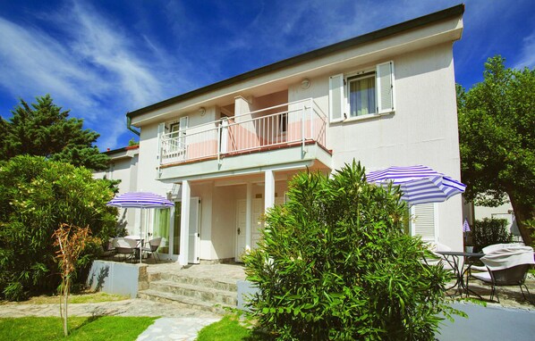 Une maison à deux étages avec un balcon, un patio couvert comprenant des tables et des chaises, et un petit jardin.