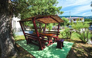 Un belvédère en bois, doté de bancs et d’une table, entouré d’arbres et surmonté d’un tapis vert posé sur le sol.