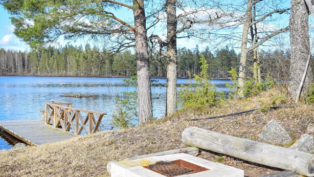 En träbrygga vid sjön med en eldstad av betong och en bänk.