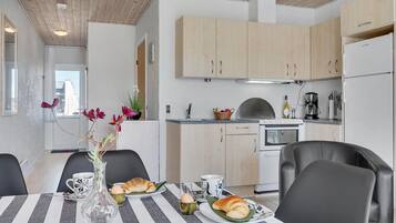 Et moderne køkken med lyse træskabe, en ovn i rustfrit stål og et køleskab. Der er et spisebord med dug i striber, stole og en vase med blomster.