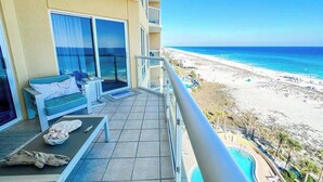 A balcony with a view of the beach, a pool, and a lounge area with a sofa and a table.