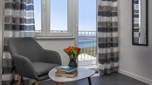 Una habitación con un sillón gris, una pequeña mesa blanca con libros, un jarrón con flores rojas y vistas al mar a través de una ventana con cortinas a rayas.